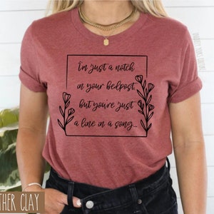 May include: A maroon t-shirt with a black graphic that reads "I'm just a notch in your bedpost but you're just a line in a song..." with floral accents.