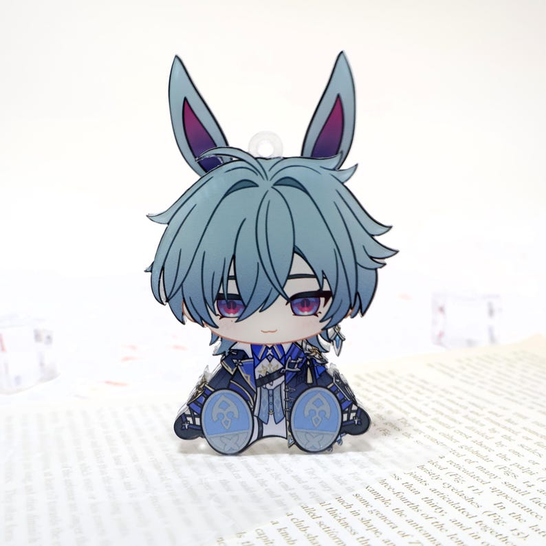 Puede incluir: Un charm de acr&iacute;lico estilo caricatura que presenta un personaje con cabello azul y orejas de conejo. El personaje lleva gafas y un atuendo detallado con detalles azules y blancos. El charm tiene un peque&ntilde;o lazo en la parte superior.
