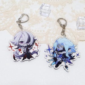 May include: Two acrylic keyrings with anime-style character designs. One keyring features a character with white and black hair, wearing a dark outfit. The other keyring has a character with blue hair and a blue and black outfit. Both have silver key rings.