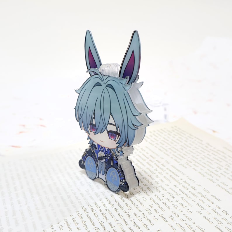 Puede incluir: Una figura de acr&iacute;lico estilo caricatura de un personaje con cabello azul y orejas de conejo. La figura tiene ojos morados y viste un atuendo detallado. La figura est&aacute; de pie sobre un libro, con el texto del libro visible.