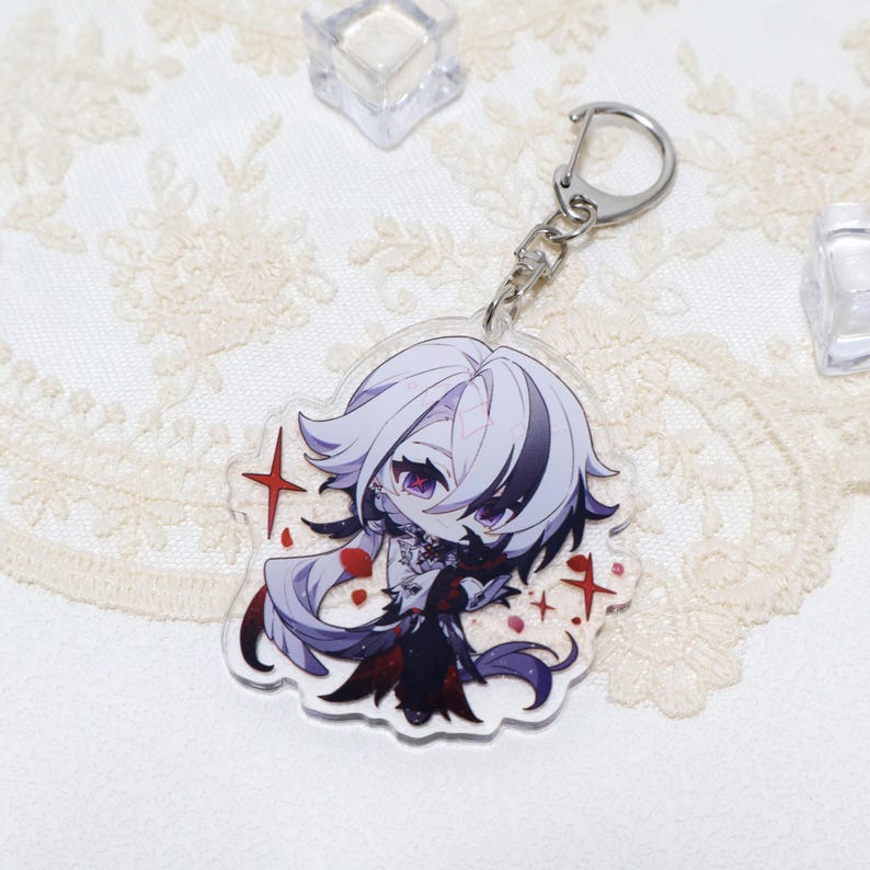 May include: A clear acrylic keychain featuring a chibi anime character with white and black hair, purple eyes, and a black outfit. The keychain has a silver metal ring and clasp. Red star accents are around the character.