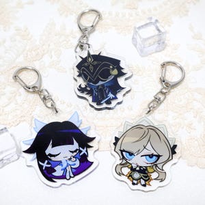May include: Clear acrylic keychains featuring anime-style character designs. Each keychain has a silver-colored metal clasp. The characters have detailed facial features and are dressed in various outfits. The keychains are on a white lace surface.