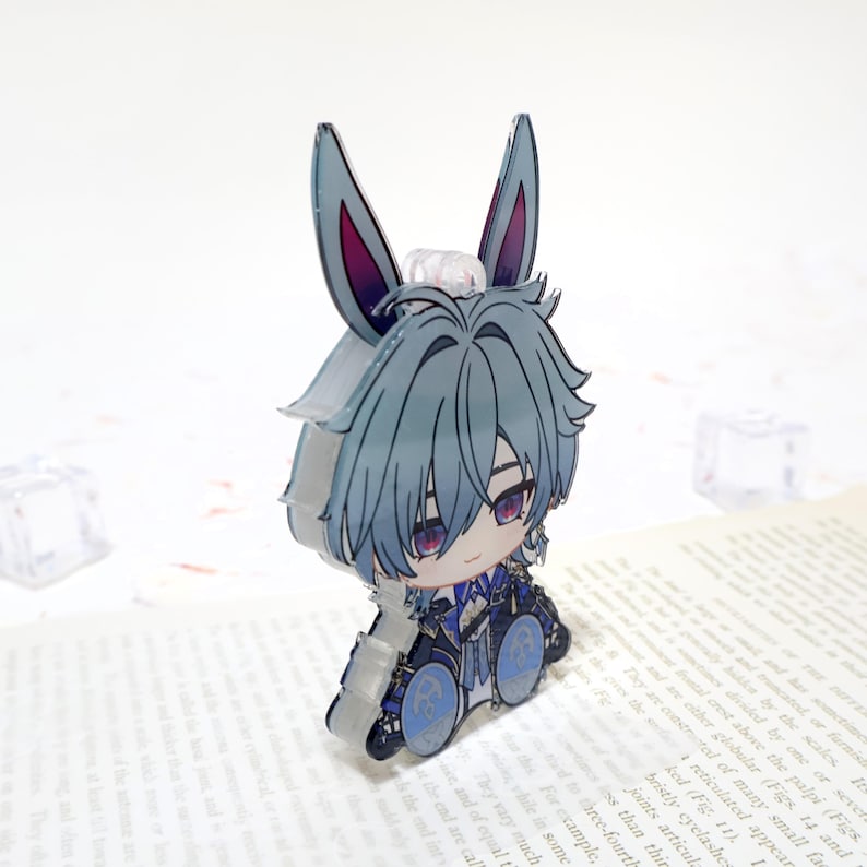 Puede incluir: Un charm de acr&iacute;lico transparente con un personaje de dibujos animados con cabello azul, ojos morados y orejas de conejo. El personaje lleva un atuendo detallado con detalles azules y negros. El charm est&aacute; sobre un libro.