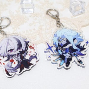 May include: Two anime-style character keychains. One keychain features a character with white and black hair, wearing a dark outfit. The other keychain has a character with blue hair and a blue and black outfit. Both have silver key rings.