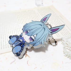 Puede incluir: Un charm de acr&iacute;lico de estilo caricatura que presenta un personaje con cabello azul claro y orejas de conejo. El charm tiene una cadena plateada y un dise&ntilde;o detallado con acentos azules y morados. El personaje est&aacute; acostado.