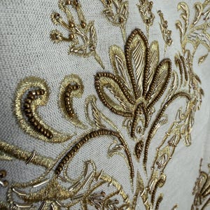 May include: A light beige fabric featuring intricate gold embroidery and metallic beadwork. The design consists of stylized floral and scrolling leaf patterns created with gold thread, small bronze-colored beads, and thin metallic tubes.