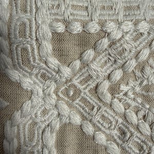 Op de afbeelding: Een close-up van beige linnen stof met ingewikkeld wit borduurwerk. Het ontwerp bestaat uit verhoogde, dikke witte draadpatronen die geometrische vormen vormen, waaronder gevlochten lijnen, rechthoekige randen en kleine cirkelvormige knopen.