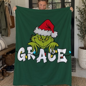 May include: A dark green fleece blanket featuring a graphic of a green character wearing a red and white Santa hat. Below the character, the name GRACE is printed in large white letters, intertwined with a string of colorful Christmas lights.