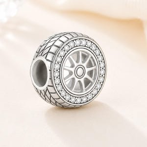 Könnte beinhalten: Ein silberner Charm in Form eines Autorads. Der Reifen hat ein strukturiertes Muster, und das Rad ist mit Speichen und einer zentralen Nabe versehen. Der Rand ist mit kleinen, klaren Edelsteinen verziert, die dem Design einen Hauch von Glanz verleihen.