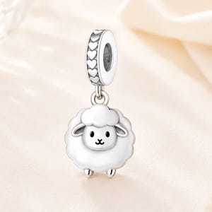 Puede incluir: Un charm de plata con un cordero blanco con un cuerpo esponjoso y una cara sonriente. El charm tiene un lazo adornado con recortes en forma de corazón para sujetarlo a una pulsera o collar. El charm de oveja es un accesorio lindo y caprichoso.