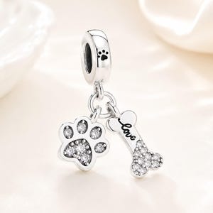 May include: A silver charm with a paw print and a bone with the word "love". The charm has a paw print on the top and is decorated with clear crystals. The charm is designed for a bracelet or necklace.