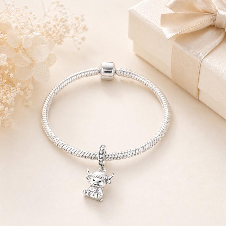 Pode incluir: Uma pulseira de prata com um berloque com uma pequena vaca Highland. O berloque est&aacute; suspenso de um conector de contas. A pulseira &eacute; exibida em uma superf&iacute;cie clara, com uma caixa de presente e flores ao fundo. Circunfer&ecirc;ncia de aproximadamente 19 cm.