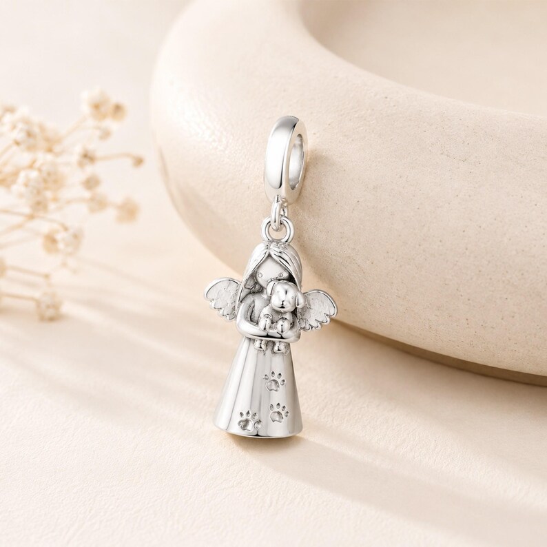 May include: A silver charm depicting an angel cradling a small dog. The angel has detailed wings and a dress decorated with paw prints. The charm is designed to be attached to a bracelet or necklace.