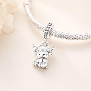 Pode incluir: Um berloque de prata com um design de vaca das Terras Altas. O berloque est&aacute; pendurado em uma pulseira de prata. A vaca tem um rosto sorridente, pequenos chifres e p&ecirc;lo detalhado. A argola do berloque &eacute; decorada com cora&ccedil;&otilde;es.