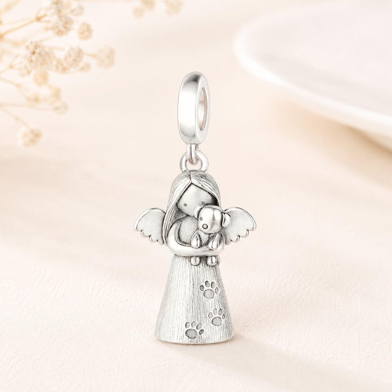 May include: A silver charm depicting an angel embracing a small dog. The angel has wings and a dress decorated with paw prints. The charm includes a loop for attaching to a bracelet or necklace. The charm is made of a shiny, metallic material.