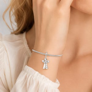 May include: A silver charm bracelet featuring an angel pendant. The angel charm is adorned with clear crystals and has detailed wings. The bracelet is displayed on a wrist, with a blurred background.
