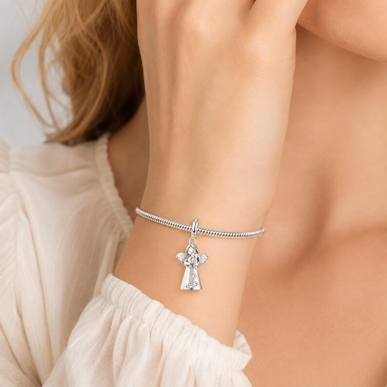 May include: A silver charm bracelet featuring an angel pendant. The angel charm is adorned with clear crystals and has detailed wings. The bracelet is displayed on a wrist, with a blurred background.