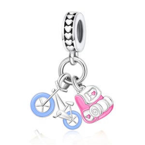 Puede incluir: Un charm de plata con un anillo de corazones recortados, del que cuelgan dos dijes: una bicicleta plateada con ruedas azul claro y una mochila rosa y blanca con pequeños detalles de corazones rosas en el frente y el lateral.