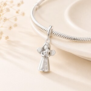May include: A silver charm depicting an angel cradling a small dog. The angel has detailed wings and a dress decorated with paw prints. The charm is suspended from a silver bracelet, set against a soft, neutral background.