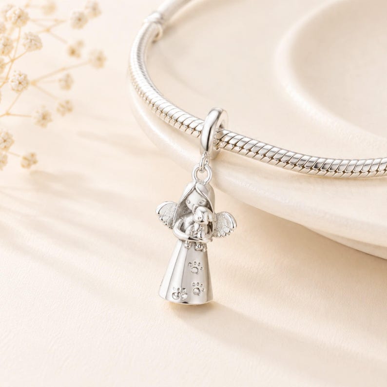 May include: A silver charm depicting an angel cradling a small dog. The angel has detailed wings and a dress decorated with paw prints. The charm is suspended from a silver bracelet, set against a soft, neutral background.