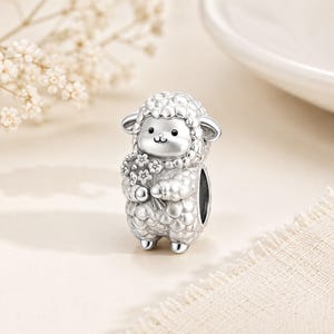 Puede incluir: Un charm de plata con forma de oveja, sosteniendo un ramo de flores. La oveja tiene un pelaje texturizado y una cara sonriente. El charm está diseñado para ser añadido a una pulsera o collar.