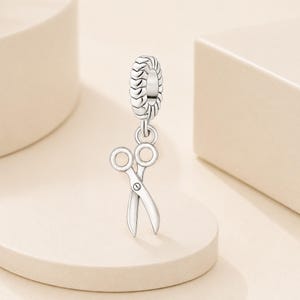 May include: A silver charm in the shape of a pair of scissors, featuring a textured, ridged cylindrical bead at the top for attachment. The scissors are depicted in an open position with two circular finger loops and two pointed blades.