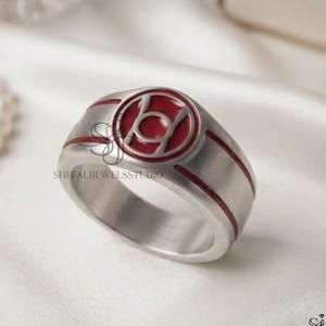 May include: A silver-toned metal ring featuring a circular emblem with a red enamel inlay. The emblem displays a stylized symbol resembling a circle with a line through it. Two thin red enamel lines wrap horizontally around the band.