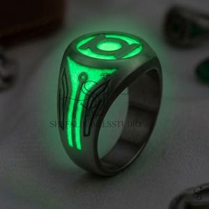 Puede incluir: Un anillo de metal oscuro con un emblema circular que presenta un símbolo de linterna verde brillante. La banda muestra intrincadas líneas negras grabadas que se extienden desde el icono central, creando un diseño de alto contraste que brilla.