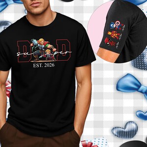 Può includere: T-shirt nera con una grafica di Thor e un bambino, con le parole "Super Dad" e "EST. 2026" in rosso e bianco. La manica presenta immagini di cartoni animati di Capitan America, Iron Man e Deadpool.