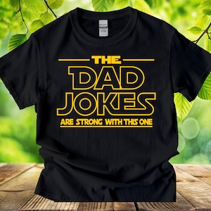 Puede incluir: Camiseta negra con texto amarillo que dice "THE DAD JOKES ARE STRONG WITH THIS ONE". El texto está en negrita y en un tipo de letra en bloque. La camiseta está hecha de un material suave y es adecuada para uso casual.