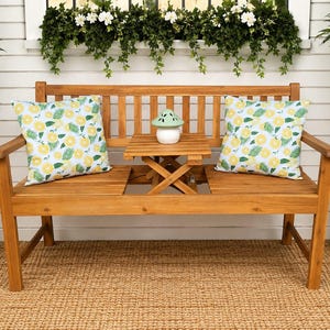 May include: A wooden bench with a slatted back and seat, featuring a built-in central table. Two square pillows with a yellow lemon and green leaf pattern rest on the seat. A small, light green mushroom-shaped ceramic object sits on the table.