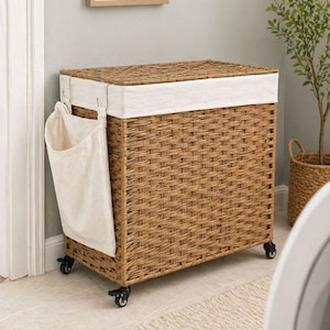 May include: A rectangular woven laundry hamper with a light brown finish, featuring a white fabric liner and a side pocket. The hamper sits on small black wheels for easy mobility. A neutral-toned rug is in front of the hamper.