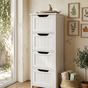 May include: A tall, white wooden storage cabinet with four drawers, each featuring a black handle. The cabinet is set against a neutral-toned wall, with framed botanical prints and a woven basket holding rolled towels. A small potted plant is also visible.