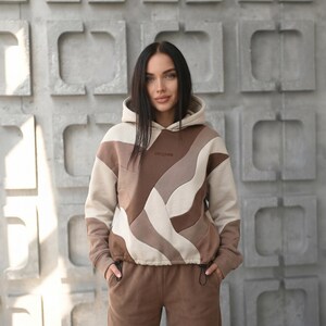 Op de afbeelding: Een beige, bruine en crèmekleurige hoodie met een taille met trekkoord en een bijpassende joggingbroek. De hoodie heeft een uniek golvend patroon en het woord "PRESTIGE" op de borst.