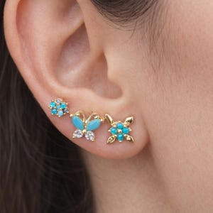 Könnte beinhalten: Goldfarbene Ohrringe mit drei Designs: eine Blume, ein Schmetterling und eine quadratische Blume. Jeder Ohrring ist mit türkisfarbenen und klaren Edelsteinen verziert. Ein zartes und auffälliges Accessoire.