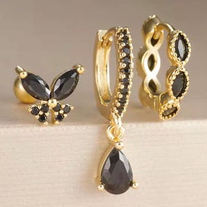 Könnte beinhalten: Ein Set aus drei goldfarbenen Ohrringen. Einer ist ein Schmetterlingsstecker mit schwarzen Edelsteinen, ein weiterer ist ein Reifen mit schwarzen Edelsteinen, und der dritte ist ein Reifen mit einem schwarzen Tropfenanhänger.