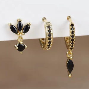 Könnte beinhalten: Drei goldfarbene Ohrringe mit schwarzen Edelsteinen. Ein Ohrring hat eine Anordnung aus drei schwarzen Steinen in Marquiseform mit einem runden, hängenden Stein. Die anderen beiden sind Creolen, eine mit schwarzen Steinen und die andere mit einem hängenden schwarzen Stein.