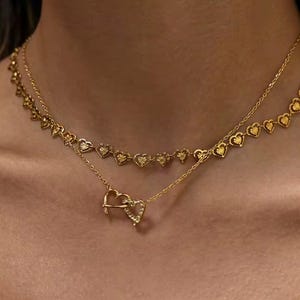 Könnte beinhalten: Ein geschichtetes goldfarbenes Halskettenset bestehend aus zwei verschiedenen Ketten. Die obere Kette besteht aus einer Reihe kleiner, miteinander verbundener herzförmiger Glieder. Die untere Kette hat einen Anhänger aus zwei ineinandergreifenden Herzen.