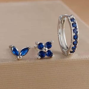Könnte beinhalten: Ein Set aus drei silbernen Ohrringen. Einer ist ein Creolen-Ohrring mit blauen Edelsteinen, ein weiterer ist ein Ohrstecker mit einem vierblättrigen Blumenmuster und der dritte ist ein Ohrstecker mit einem Blattmuster.