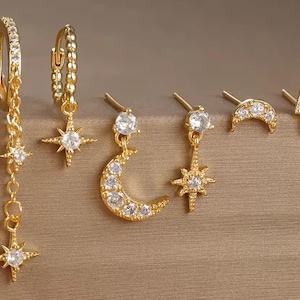 Op de afbeelding: Goudkleurige oorbellen met hemelse ontwerpen. De set bevat oorringen met sterhangers, oorknopjes in de vorm van sterren, manen en een bliksemschicht. Elke oorbel is versierd met heldere edelstenen.