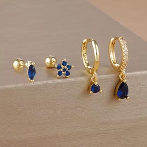 Könnte beinhalten: Eine Sammlung goldfarbener Ohrringe mit blauen Edelsteinakzenten. Das Set umfasst Ohrstecker mit Tropfen- und Blumenmuster sowie Creolen mit einem Tropfenanhänger. Die Ohrringe werden auf einer neutralen Oberfläche präsentiert.