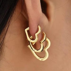 Könnte beinhalten: Goldfarbene herzförmige Ohrringe. Die Ohrringe bestehen aus glänzendem Metall und haben ein mehrschichtiges Design. Die Ohrringe werden am Ohr getragen und sind auf der Haut sichtbar.