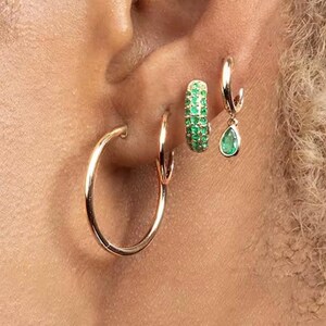 Può includere: Primo piano di un orecchio adornato da più orecchini. Un grande orecchino a cerchio dorato è nel primo piercing. Il secondo piercing ha un cerchio d'oro più piccolo. Il terzo piercing ha un piccolo cerchio d'oro con una goccia di smeraldo verde. Il quarto piercing ha un piccolo cerchio d'oro con smeraldi verdi.