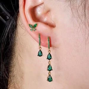 Könnte beinhalten: Ein Set aus goldfarbenen Ohrringen, bestehend aus einem schmetterlingsförmigen Stecker mit grünen Steinen, einem kleinen Creolen-Ohrring mit einem tropfenförmigen grünen Stein und einem längeren Creolen-Ohrring mit drei hängenden grünen Tropfen.