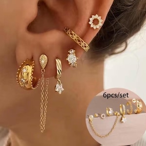 Könnte beinhalten: Eine Sammlung goldfarbener Ohrringe, darunter Creolen, Ohrstecker und Hängeohrringe, einige verziert mit klaren Edelsteinen und sternförmigen Anhängern. Das Set bietet eine Vielzahl von Designs für mehrere Ohrlöcher mit verschiedenen Texturen und Formen.