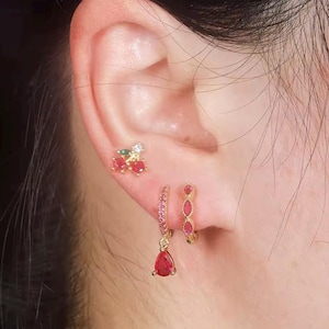 Könnte beinhalten: Goldfarbene Ohrringe mit roten und grünen Edelsteinen. Die Ohrringe umfassen einen Kirsch-Ohrstecker, einen kleinen Reifen mit rosa Steinen und einen tropfenförmigen Ohrhänger mit einem roten Stein. Der Schmuck wird an einem Ohr getragen.