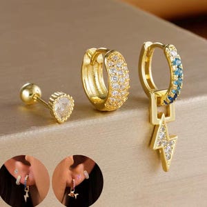 Könnte beinhalten: Eine Sammlung goldfarbener Ohrringe. Ein Ohrring ist ein Stecker mit einem klaren, facettierten Stein. Ein weiterer ist ein kleiner Reifen, der mit klaren Steinen bedeckt ist. Der dritte Ohrring ist ein Reifen mit blauen und klaren Steinen und einem Blitz-Anhänger.