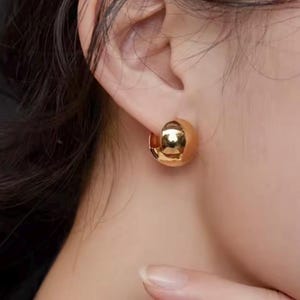 Könnte beinhalten: Nahaufnahme eines goldfarbenen Halbkreis-Ohrrings. Der Ohrring hat ein abgerundetes, poliertes Design, das das Licht reflektiert. Der Schmuck wird am Ohr einer Person mit dunklem Haar getragen.