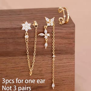 Könnte beinhalten: Ein Set aus drei goldfarbenen Ohrringen für ein Ohr, bestehend aus einem Blumenstecker mit Kette, einem Schmetterlingsstecker mit hängender Kette und einer gedrehten Ohrklemme. Jedes Teil ist mit kleinen klaren Steinen besetzt. Text: 3pcs for one ear Not 3 pairs.