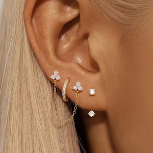 Può includere: Una collezione di orecchini color oro con scintillanti pietre di zirconia cubica. Il set include orecchini a lobo, un orecchino a cerchio e orecchini collegati da una delicata catena. Gli orecchini sono indossati su un orecchio.